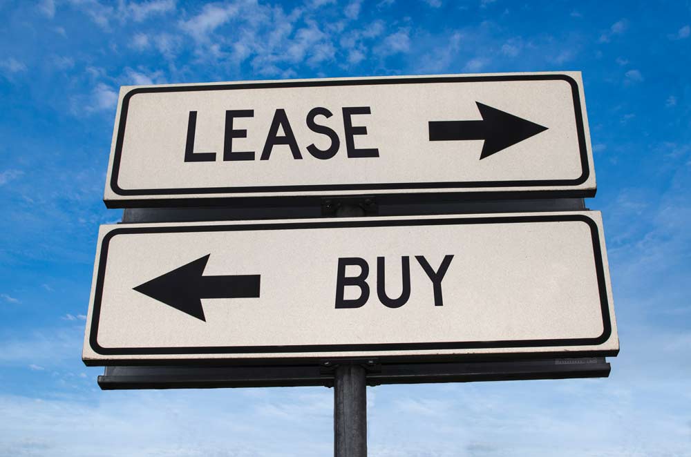 Two street signs against a blue sky; the top sign points right and says "LEASE," while the bottom sign points left and says "BUY," visually representing the classic leasing vs buying business decision.