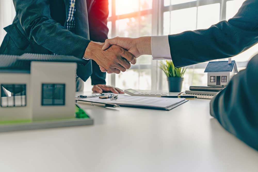 Two people in business attire shake hands across a desk with house models, documents, keys, and a plant, suggesting an office real estate deal focused on buying commercial properties or discussing investment strategies.