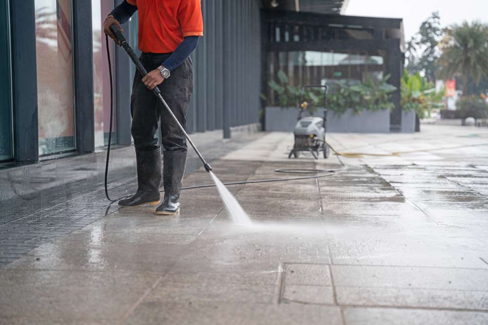 A person in rubber boots and an orange shirt uses a power washer to clean the wet, tiled outdoor pavement area near a building—demonstrating effective property management. A pressure washer machine is visible in the background.